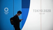 Ein Mann mit Mundschutz geht im Bahnhof Shinjuku in Tokio an einer Werbefläche für die Olympischen Spiele 2020 in Tokio vorbei. © picture alliance/Stanislav Kogiku/SOPA Images via ZUMA Wire/dpa Foto: Stanislav Kogiku