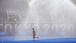Eine deutsche Spielerin läuft vor Spielbeginn in Tokio durch den Sprühregen der Platzbewässerung. © dpa picture alliance Foto: Michael Kappeler