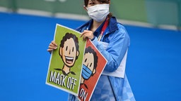 Eine Olympia-Mitarbeiterin hält Hinweisschilder zum Maskentragen hoch. © imago images/Sven Simon 