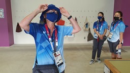 Drei freiwillige Helferinnen bei den Olympischen Spielen stehen gemeinsam in einem Raum, alle tragen Mund-Nasen-Schutz und offizielle blaue Helfer-T-Shirts. © ARD Foto: Julia Linn